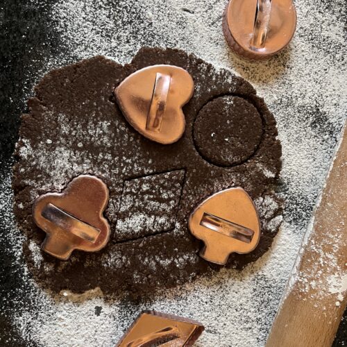 Overhead view of dark chocolate Brunsli dough rolled flat on a flour-dusted surface with copper cookie cutters in heart, circle, clover, and other shapes pressed into the dough.