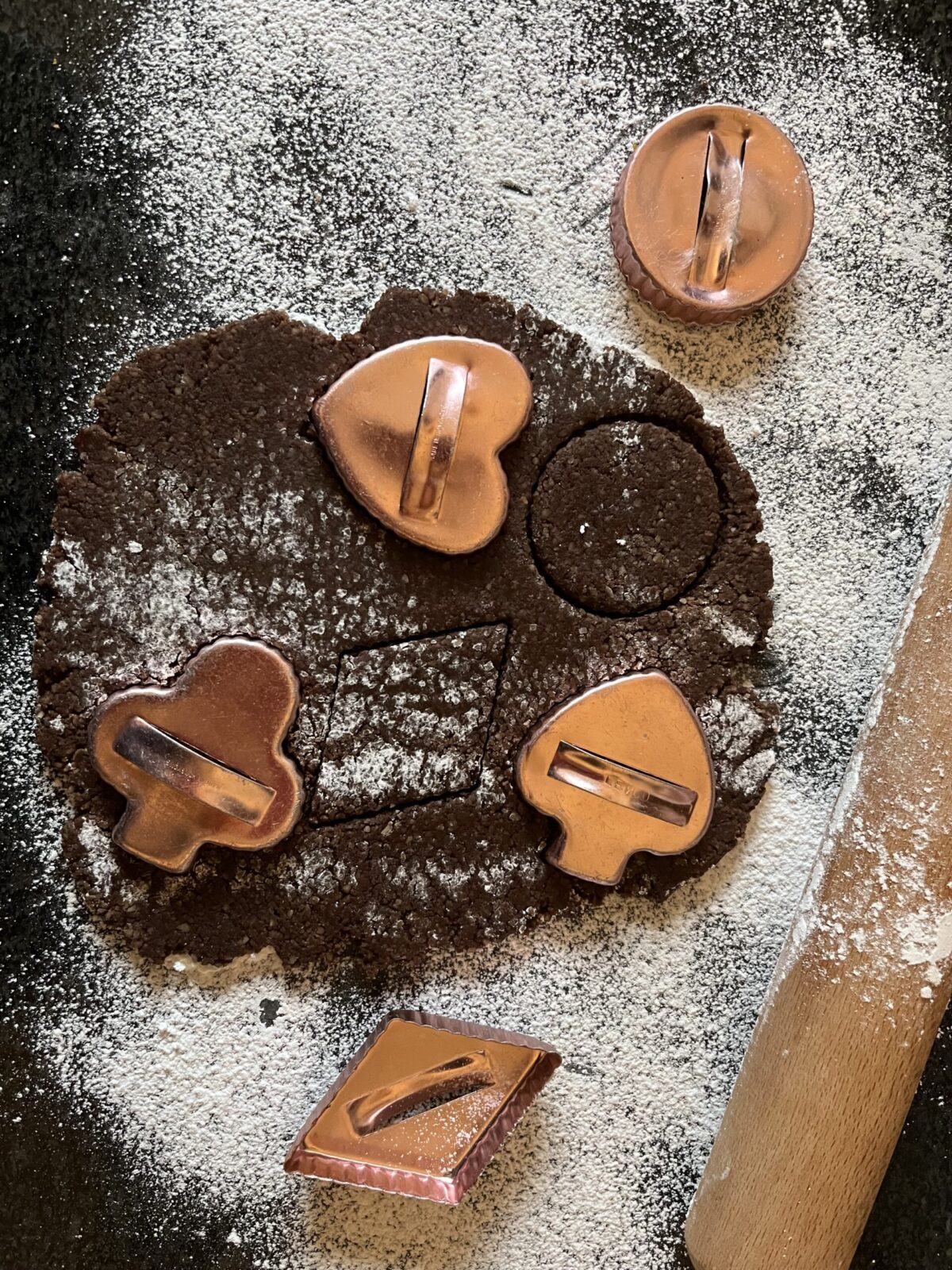 Overhead view of dark chocolate Brunsli dough rolled flat on a flour-dusted surface with copper cookie cutters in heart, circle, clover, and other shapes pressed into the dough.