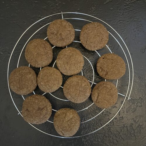 Approximately eleven baked round ginger cookies cooling on a circular wire rack on a dark slate surface, their surfaces golden-brown and lightly crackled.