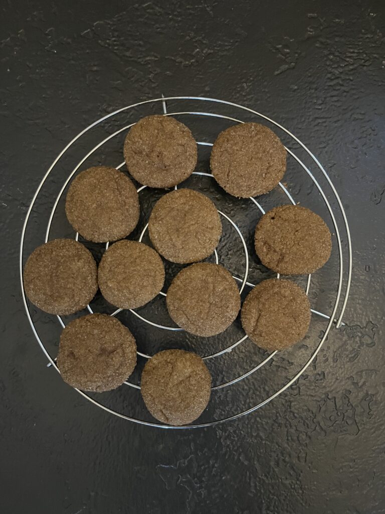 Approximately eleven baked round ginger cookies cooling on a circular wire rack on a dark slate surface, their surfaces golden-brown and lightly crackled.