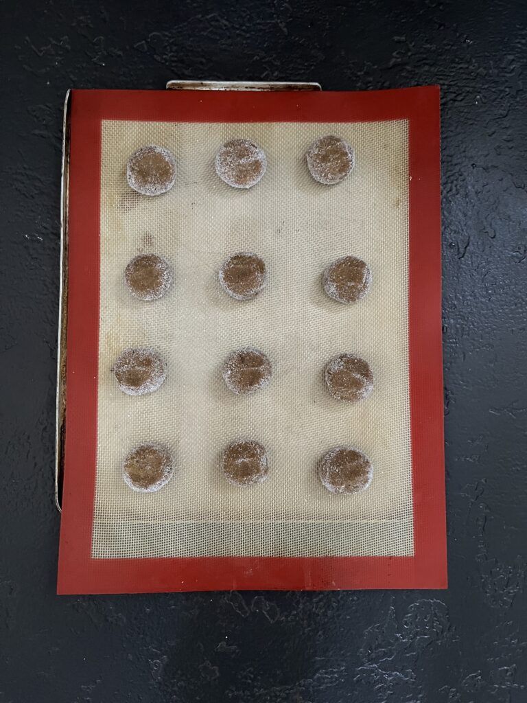 Fifteen small round balls of brown ginger cookie dough arranged in an even grid on a silicone baking mat, ready for the oven.