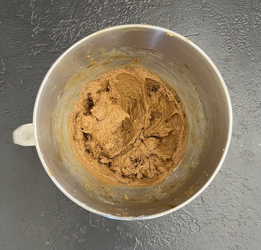 Overhead view of a stainless-steel stand-mixer bowl containing thick, smooth brown ginger cookie dough on a dark slate surface.