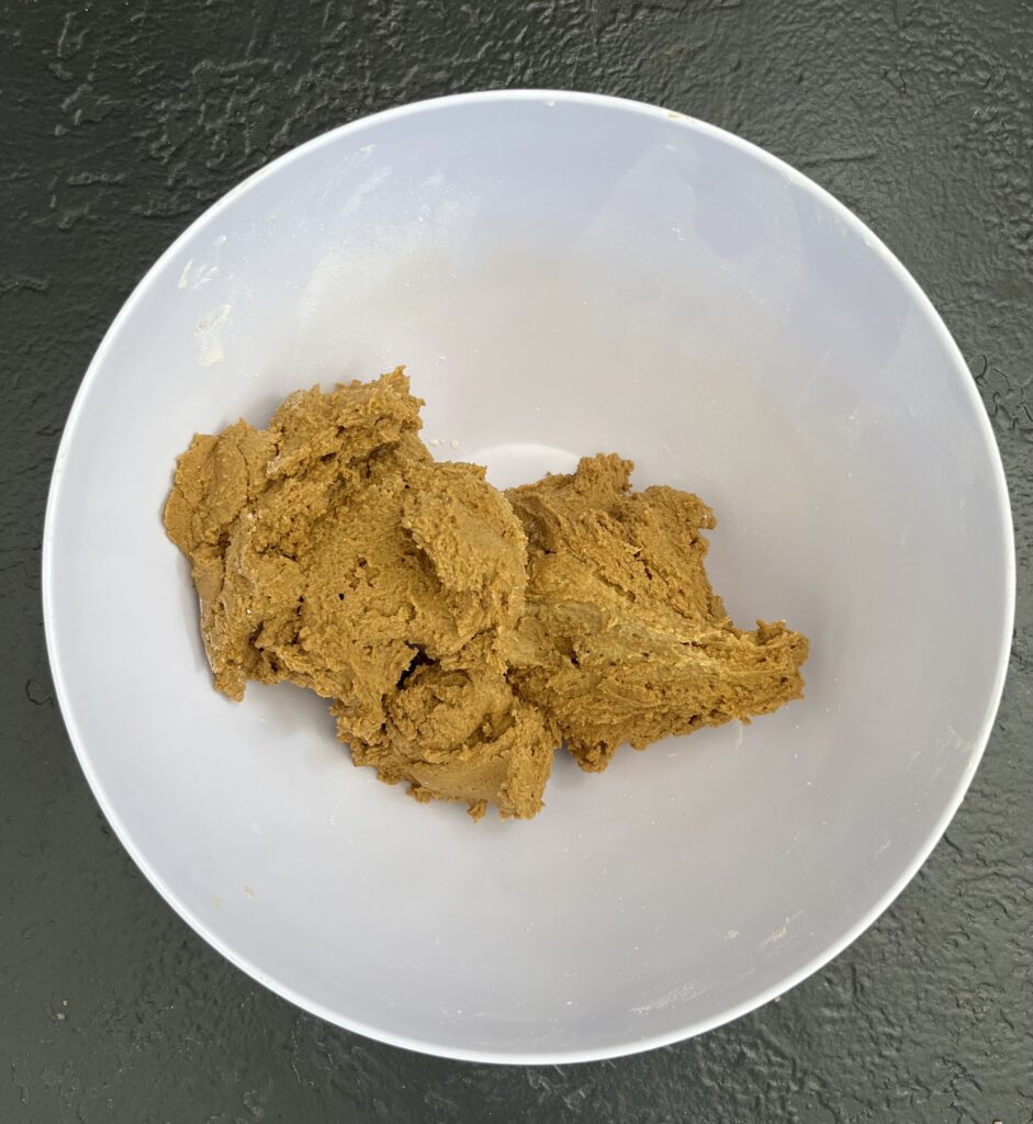 Overhead view of a white bowl containing a rough mound of golden-brown ginger cookie dough on a dark slate surface.