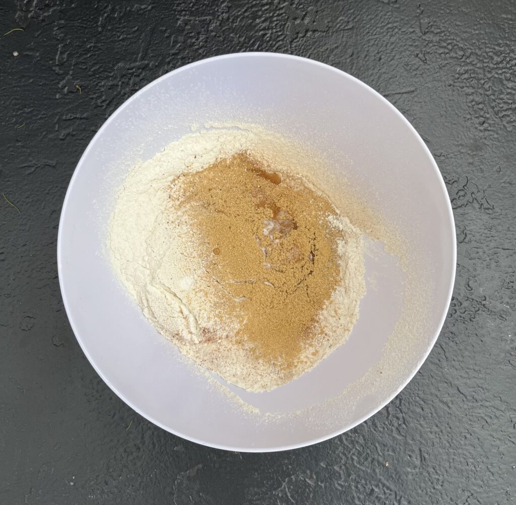 Overhead view of a white mixing bowl containing white flour with a mound of golden-brown ground ginger piled in the center, on a dark slate surface.