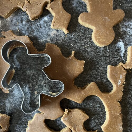 Overhead close-up of rolled-out gingerbread dough on a flour-dusted dark surface with a gingerbread man cookie cutter and several cut-out shapes.