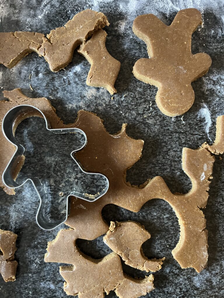 Overhead close-up of rolled-out gingerbread dough on a flour-dusted dark surface with a gingerbread man cookie cutter and several cut-out shapes.