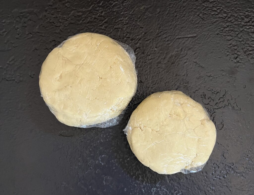 Two round discs of pale kolaczki cookie dough wrapped in clear plastic wrap on a dark slate surface, flattened and ready for chilling.
