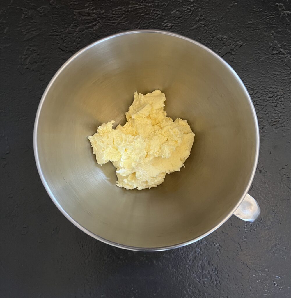 Overhead view of a stainless-steel stand-mixer bowl containing pale yellow creamed butter and cream cheese on a dark slate surface.