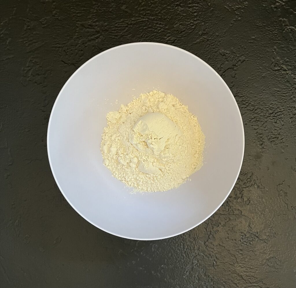 Overhead view of a large white mixing bowl containing flour and dry ingredients on a dark slate surface.