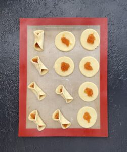 Overhead view of a silicone baking mat showing Polish kolaczki in progress, round dough circles with orange fruit filling being pinched into traditional folded shapes.