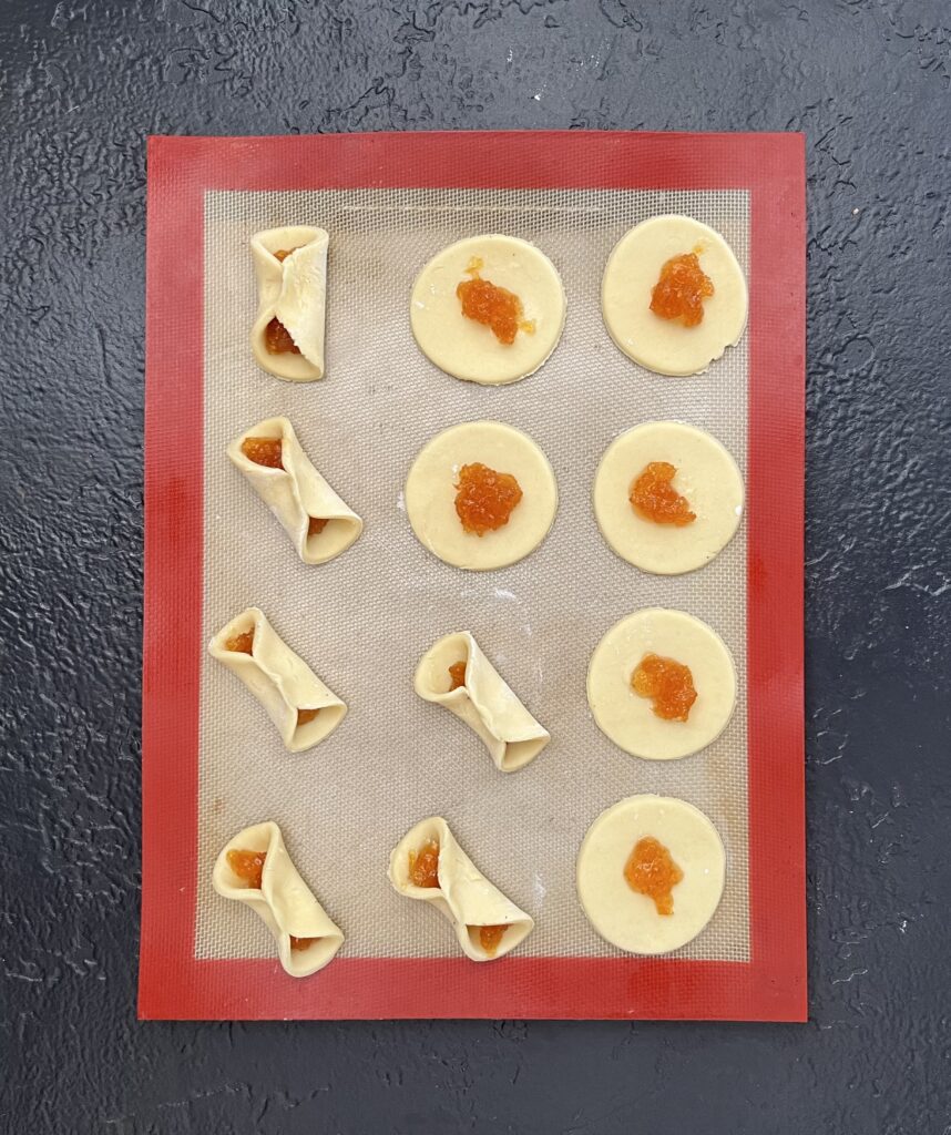 Overhead view of a silicone baking mat showing Polish kolaczki in progress — round dough circles with orange fruit filling being pinched into traditional folded shapes.
