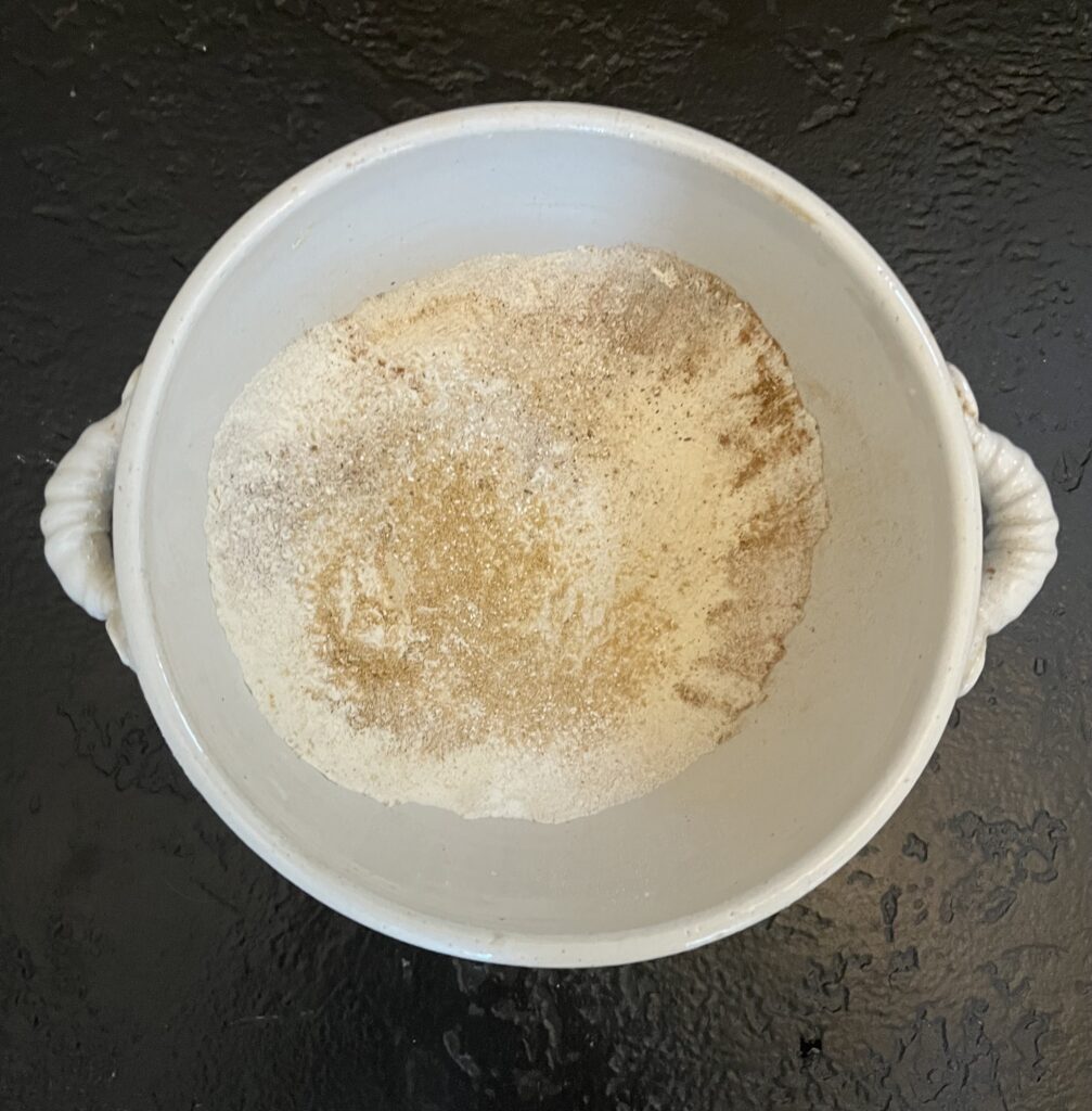 Overhead view of a white mixing bowl containing flour and ground spices swirled together on a dark slate surface.