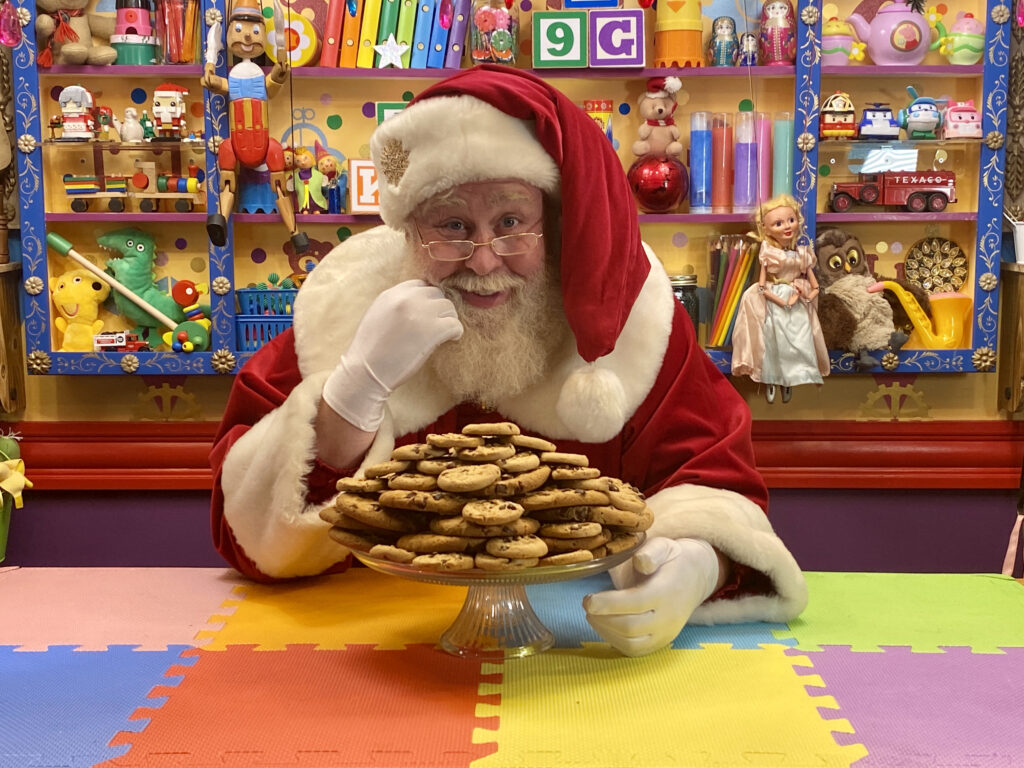 Santa Claus sitting at a colourful puzzle-mat table in a toy-filled workshop, resting his chin on one gloved hand beside a glass pedestal plate stacked high with chocolate chip cookies.