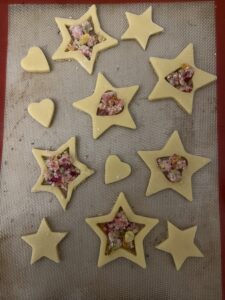 Unbaked star and heart cookie cutouts on a silicone baking mat, their centers filled with crushed pink, yellow, and amber hard candy to create a stained-glass effect.