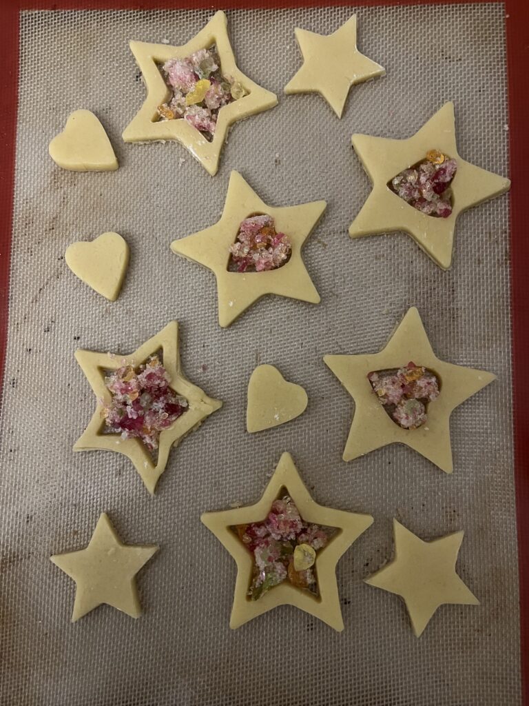 Unbaked star and heart cookie cutouts on a silicone baking mat, their centers filled with crushed pink, yellow, and amber hard candy to create a stained-glass effect.