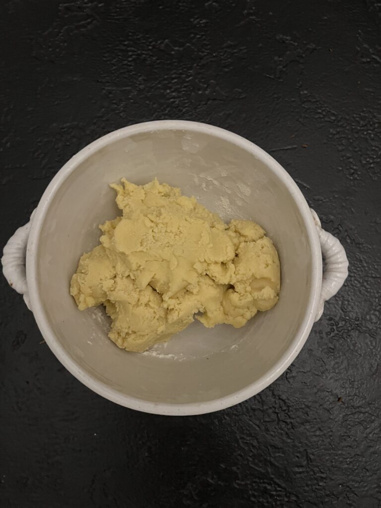 Overhead view of a white mixing bowl containing a cohesive mound of pale yellow sugar cookie dough on a dark slate surface.