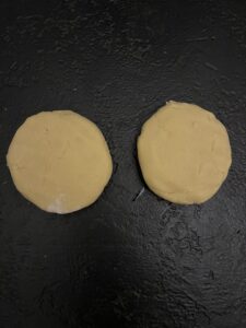 Two round, flattened discs of pale yellow cookie dough side by side on a dark surface, portioned and shaped for chilling.