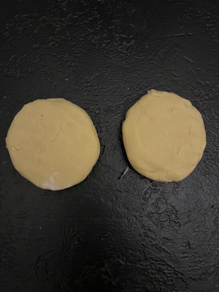 Two round, flattened discs of pale yellow cookie dough side by side on a dark surface, portioned and shaped for chilling.