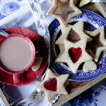 Star-shaped butter cookies with translucent candy centers in red and amber on a blue willow-pattern plate, alongside a red teacup of milky tea.