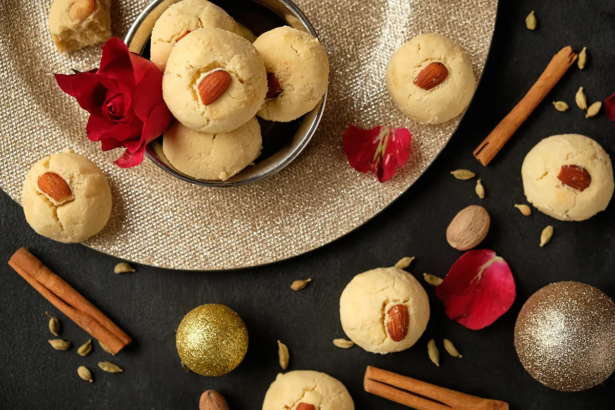 Round golden nan khatai cookies topped with whole almonds, arranged on a metallic platter with cinnamon sticks, cardamom pods, rose petals, and gold Christmas ornaments on a dark background.