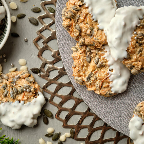 Lacy golden-brown Florentine cookies studded with pumpkin seeds and pine nuts, some partially dipped in white chocolate, arranged on a silver lattice tray with loose seeds and evergreen sprigs.