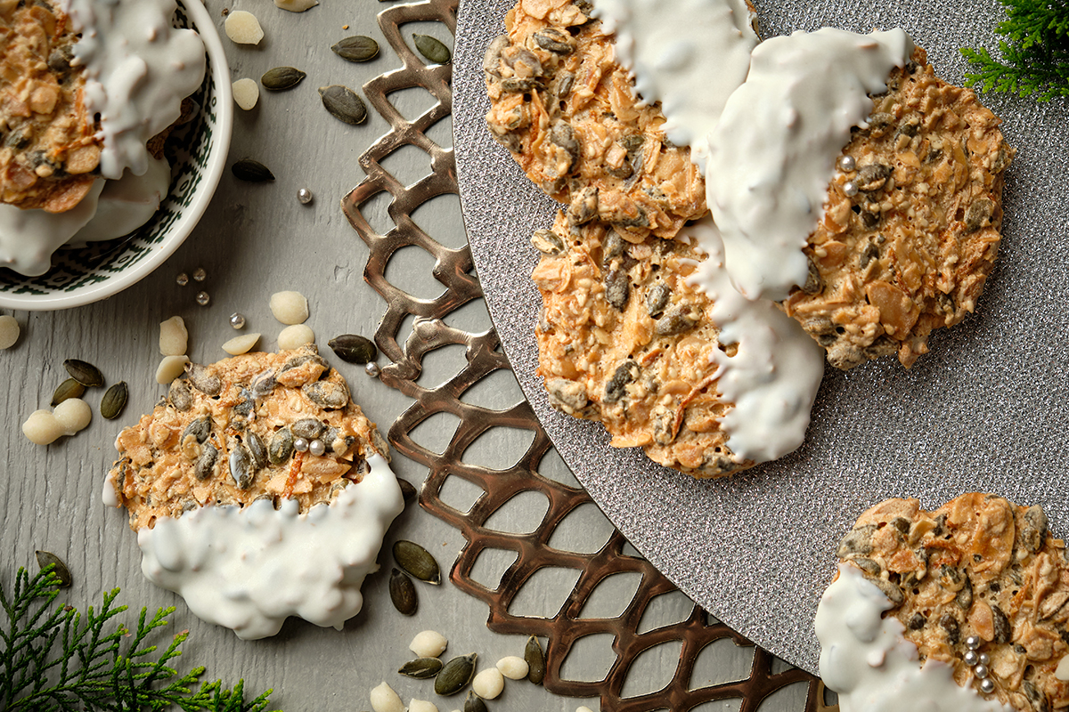 Lacy golden-brown Florentine cookies studded with pumpkin seeds and pine nuts, some partially dipped in white chocolate, arranged on a silver lattice tray with loose seeds and evergreen sprigs.