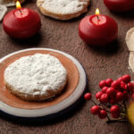 Round powdered-sugar-coated polvorones cookies on rustic terracotta plates with red candles, holly berries, and lace doilies.