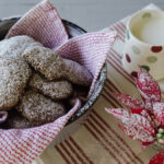 Thick dark ginger cookies dusted with powdered sugar in a dark bowl lined with red-and-white checkered cloth, with a polka-dot milk jug and red poinsettia.