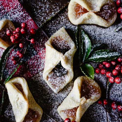 Overhead close-up of baked kolaczki cookies folded into diamond shapes, dusted with powdered sugar and garnished with fresh red berries and bay leaves on a dark surface.