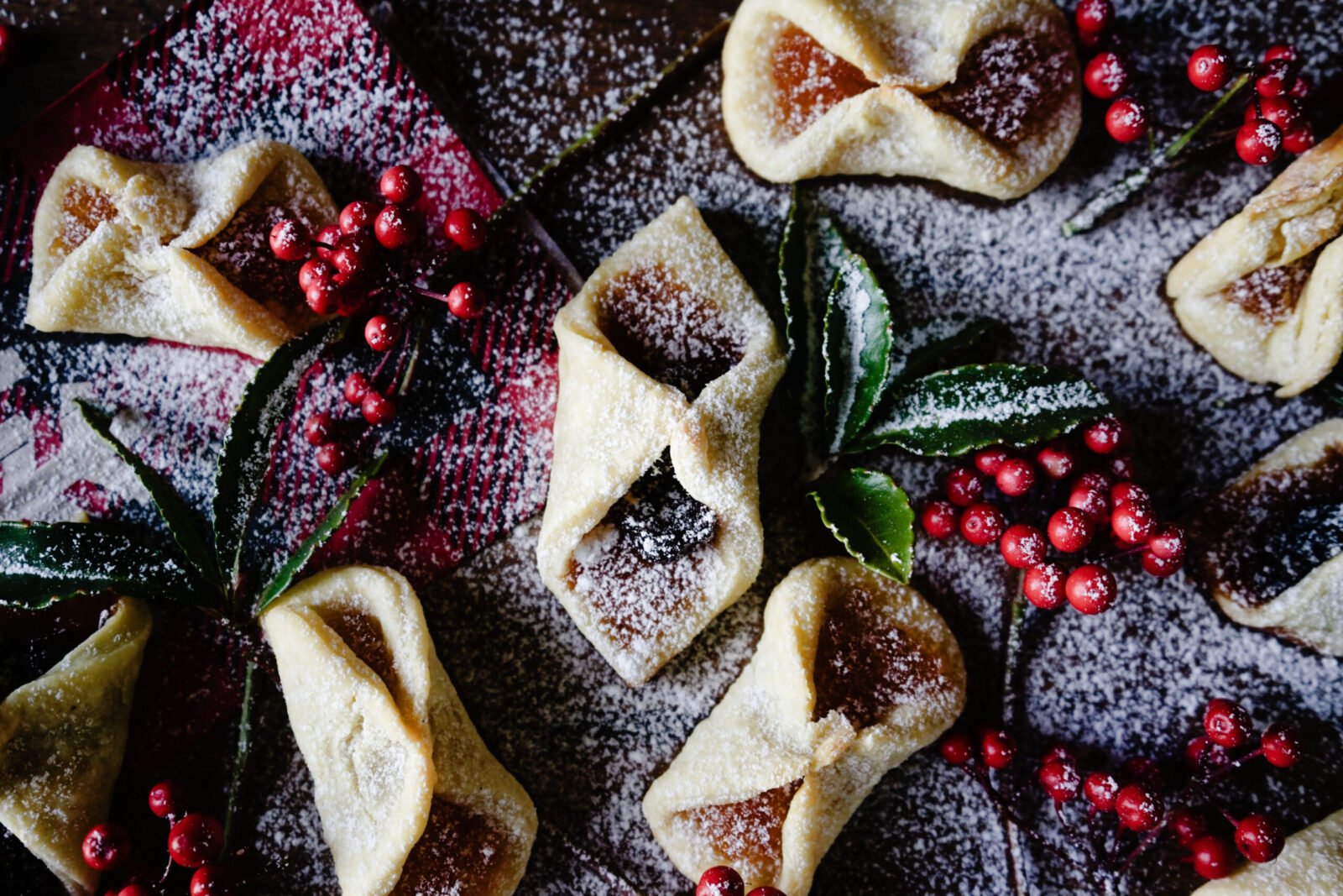 Overhead close-up of baked kolaczki cookies folded into diamond shapes, dusted with powdered sugar and garnished with fresh red berries and bay leaves on a dark surface.