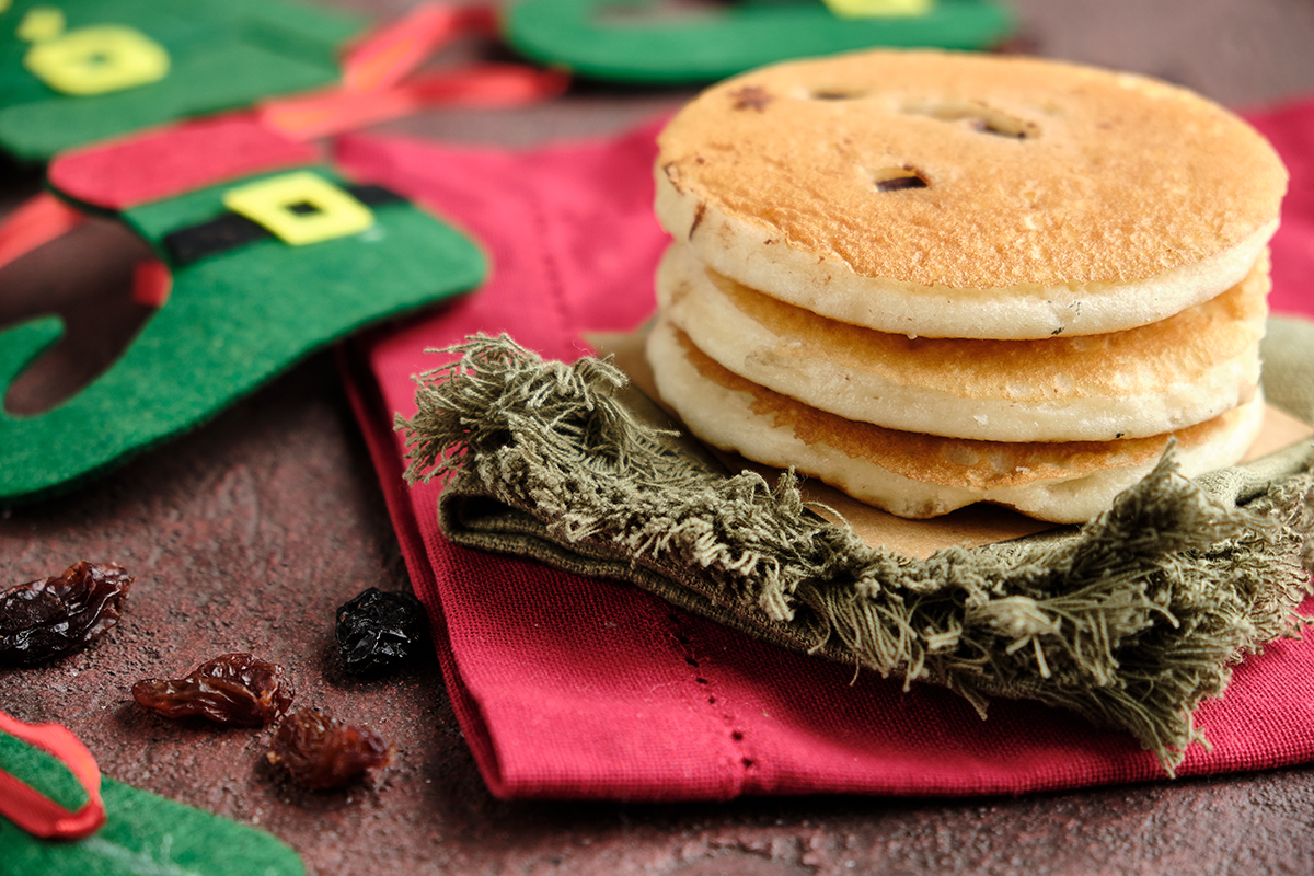A centered stack of four currant-studded Welsh cakes on green and red napkins, with felt elf advent-calendar stockings and warm bokeh lights in the background.