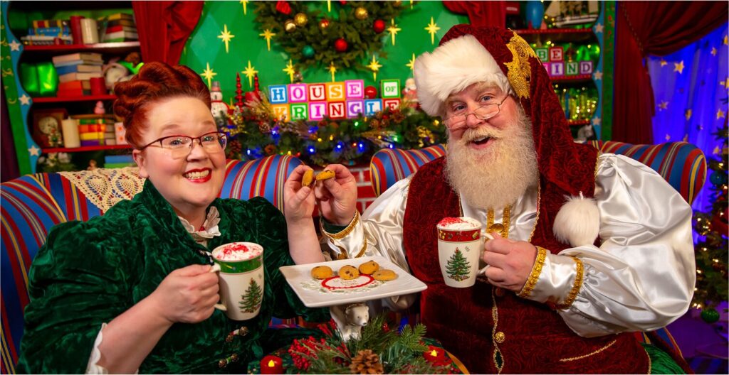 Santa Claus and Mrs. Claus sitting together on striped chairs, holding Christmas tree mugs with whipped cream and sharing a plate of cookies.